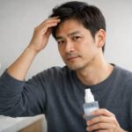 30代後半の日本人男性が自宅のベッドに腰かけておでこの生え際にそっと手を添えながら、薄毛対策や育毛剤の情報を真剣な表情で読み込んでいる横長ポートレート。部屋のやわらかな自然光が髪と表情をやさしく照らし、買う前にじっくり調べているリアルな不安と慎重さが伝わる一枚。 A Japanese man in his late thirties sits on his bed at home, gently touching his hairline while reading detailed information about hair loss care and hair growth tonics. Soft natural light fills the room and highlights his thoughtful expression, capturing the realistic concern and careful research before making a purchase.
