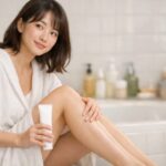 明るい室内で、20代くらいの日本人女性が片ひざを立てて座り、脚に除毛クリームをなじませたあと、なめらかな素肌をやさしく見つめている横長ポートレート / A young Japanese woman sits in a bright room with one knee up, gently applying hair removal cream to her lower leg and looking at her smooth skin with a soft, relaxed smile in a horizontal portrait