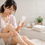 肌が弱めの女性が、自宅のリビングで脚の仕上がりを確認しながら除毛クリームでのムダ毛ケアを考えている様子 / A young Japanese woman in a bright living room gently touching her smooth legs and thinking about using depilatory cream for gentle hair removal