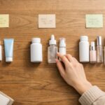 木目のテーブルの上に、左・中央・右と3列に並べられた白いサプリボトルや美容液ボトル、日焼け止めチューブが俯瞰で写り、右下には電卓、左下にはノートとペン、中央にはベージュのニットを着た女性の手が中央のボトルにそっと触れている「予算別スキンケアセット」を考えている横長フラットレイ写真｜A wide horizontal overhead flatlay photo on a wooden table showing three groups of plain supplement bottles, serum bottles and sunscreen tubes from left to right, with a calculator and notebook at the edges and a woman’s hand in a beige sweater gently adjusting one bottle in the middle group, representing budget-based self-care sets.